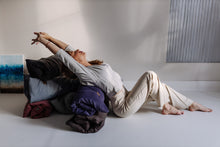 Załaduj obraz do przeglądarki galerii, Na zdjęciu kobieta w jasnym stroju wykonuje ćwiczenia rozciągające w pozycji leżącej, opierając się o stos złożonych mat do medytacji i jogi w różnych kolorach. Jasne, minimalistyczne wnętrze podkreśla atmosferę relaksu i harmonii.