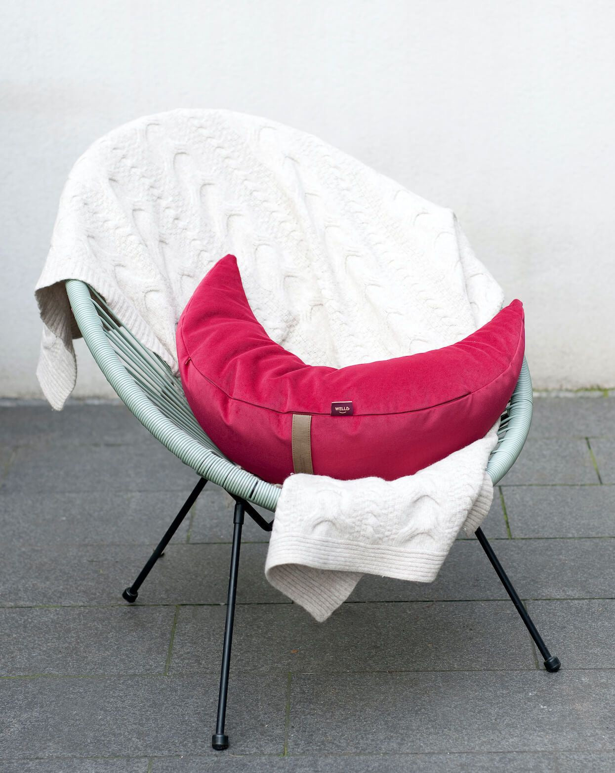 poduszka księżycowa w welurowym pokrowcu w kolorze malinowym leżąca na fotelu wiklinowym
