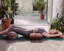 Load image into Gallery viewer, kobieta praktykująca jogę leżąc na macie z poduszką kopertową pod lędźwiami i bolsterem  gryczanym pod kolanami
