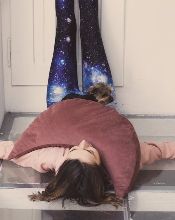 kobieta leżąca na podłodze opierając nogi na drzwiach trzymając na klatce poduszkę księżycową w kolorze ciemno różowym i małego psa